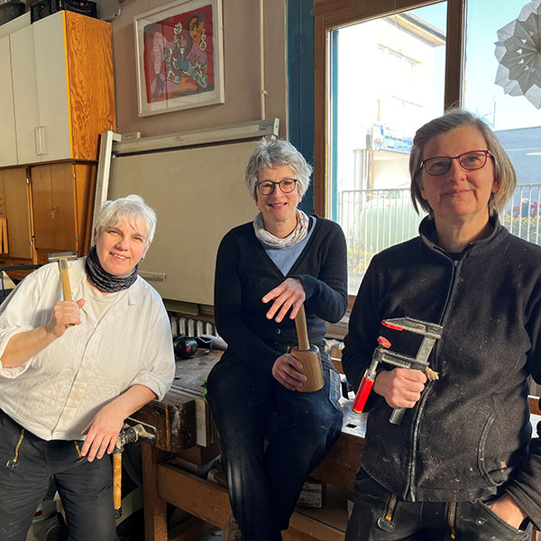 Drei Tischlerinnen lächeln in ihrer Werkstatt.